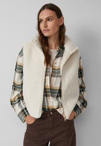 Fleece-Weste in Off-White mit hohem Kragen und Reißverschluss. Über einem karierten Hemd in Grün-, Gelb- und Grautönen getragen. Braune Hose sichtbar.