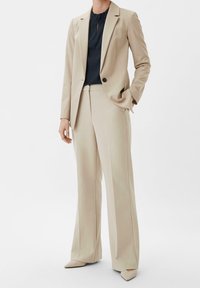 Completo tailored beige con giacca aderente e pantaloni a gamba larga. Sotto c'è un top blu navy con dettagli in pizzo sul colletto. Scarpe beige a punta.