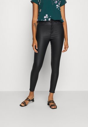 Pantalons noirs moulants taille haute avec une texture lisse, associés à un haut floral vert foncé et des talons noirs à lanières.
