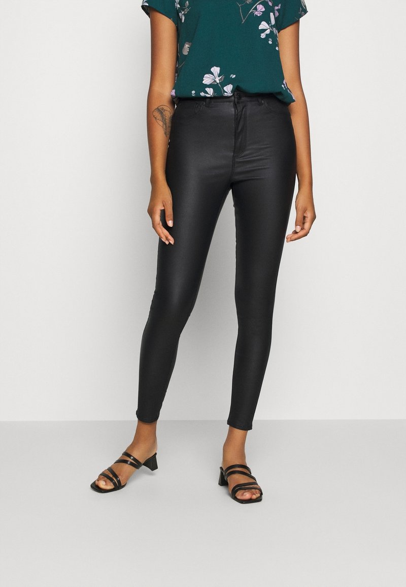 Pantalones negros, ajustados y de talle alto con una textura suave, combinados con una blusa floral de color verde oscuro y tacones negros con tiras.