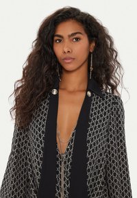 Donna con lunghi capelli ricci e scuri, che indossa un blazer a motivi nero e beige, con collane d'argento a strati e orecchini pendenti.