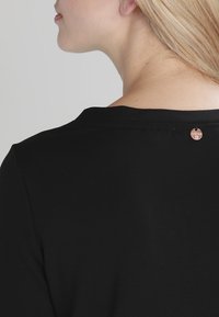 Svart skjorta med rund halsringning, korta ärmar och en liten logotagg i kopparfärg bak. Slät tygtextur.