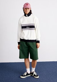 Witte half-zip sweatshirt met zwarte en grijze strepen, groene shorts, roze pet, zwarte sneakers en witte sokken. Casual athletische stijl.