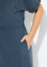 Tumma sininen mekko pehmeästä kankaasta, jossa on lyhyet hihat ja tasku vasemmalla puolella, sekä rypytys yksityiskohtana vyötäröllä.