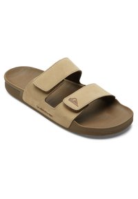 Sandales en toile beige avec deux sangles Velcro réglables, semelle incurvée et semelle texturée. Présente un logo embossé sur le côté.