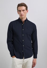 Chemise bleu marine boutonnée avec manches longues, une poche poitrine et des boutons blancs. Le tissu semble lisse sans motifs visibles. Portée avec un pantalon beige.