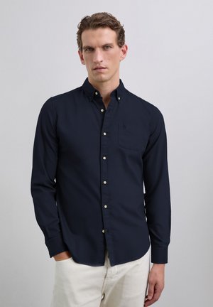 Navy button-up shirt with long sleeves, a chest pocket, and white buttons. Fabric appears smooth without visible patterns. Worn with beige pants.