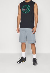 T-shirt sans manches noir avec un motif vert et rose représentant un ballon de basket. Short gris à texture lisse. Chaussures de sport noires avec des accents blancs.