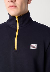 Pulover cu fermoar închis la culoare bleumarin, cu guler înalt, având un fermoar galben și o etichetă cu logo-ul Gant pe piept. Fabricat dintr-un material moale și texturat.
