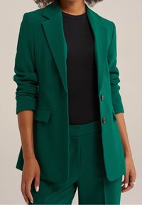 Grön kavaj i ett mjukt tyg, med slagkrage, två knappar och uppvikta ärmar.Matchad med en svart topp.