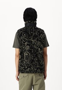 Black and cream patterned fleece vest with a high collar, featuring a textured design. Worn over a gray shirt and paired with olive-green pants.