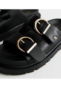 Zwarte sandalen met twee brede banden, gouden ronde gespen en een gestructureerd oppervlak. Dikke zool met een lichte plateau. Eenvoudig ontwerp.