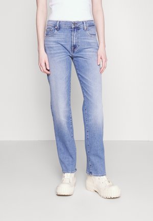 Personne portant un jean droit bleu clair, des chaussures blanches à semelle plateforme épaisses et un top blanc sans manches sur un fond clair uni.