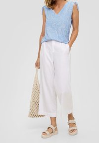 Donna che indossa un top senza maniche azzurro chiaro, pantaloni bianchi a gamba larga, sandali intrecciati beige e tiene in mano una borsa tote intrecciata beige.
