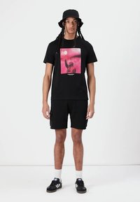 T-shirt noir avec un imprimé graphique coloré, associé à un short noir. Comprend également un chapeau seau noir et des baskets noires avec des accents blancs.