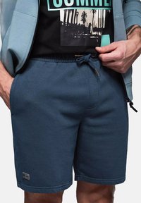 Niebieskie szorty dresowe wykonane z teksturowanej tkaniny, z elastycznym paskiem z troczkiem oraz bocznymi kieszeniami. Logo na materiale w pobliżu dołu.