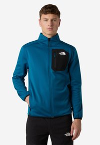 Giacca softshell in teal, con una tasca sul petto nera con zip, colletto alto e chiusura anteriore con zip. Il materiale ha una texture liscia.