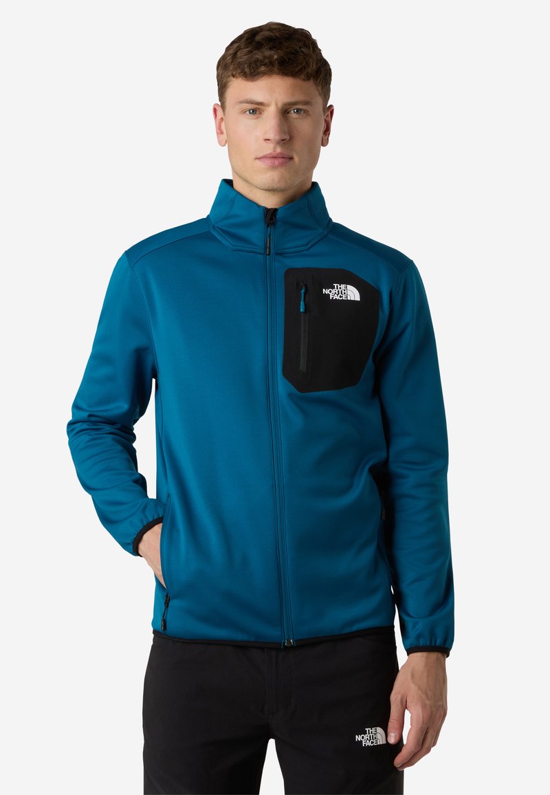 Giacca softshell in teal, con una tasca sul petto nera con zip, colletto alto e chiusura anteriore con zip. Il materiale ha una texture liscia.