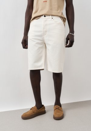 Muž nosiaci béžové tričko, biele šortky po kolená, hnedé semišové mokasíny a strieborný náramok na pravom zápästí, stojaci na sivej podlahe.