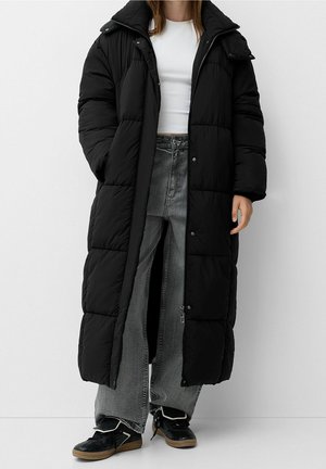 Femme portant un long manteau matelassé noir par-dessus un crop top blanc, un jean large gris et des baskets noires à semelles en gomme, debout contre un mur blanc.