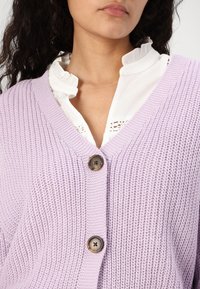 Lys lilla strikket cardigan med v-hals, med tre knapper foran, og en ribbestruktur. Underneath er det en hvit volangkrage.