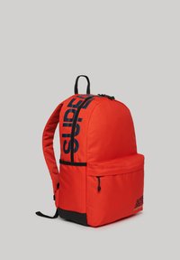 Mochila de tela roja con forma redondeada, cremallera y detalles negros, bolsillo frontal y un gran texto de logo en un lado.