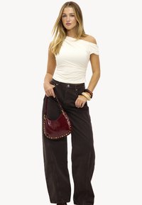 Haut blanc sans épaules, jeans large marron foncé, sac à main bordeaux brillant avec des clous dorés, et bracelets épais.