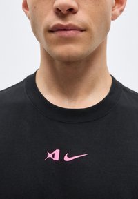 Κοντινό πλάνο ενός ατόμου που φοράει μαύρο t-shirt με δύο μικρά ροζ λογότυπα στο στήθος, συμπεριλαμβανομένου ενός παραμορφωμένου λογότυπου Nike.