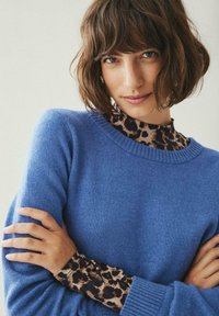 Modra pletena jopica s okroglem vratom, nošena čez leopardji potiskani lažni rolak. Tkanina se zdi mehka in lahka, z rebrastimi detajli.