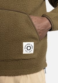 Sweatshirt en polaire marron avec une poche visible et une étiquette portant un logo circulaire, "REDEFINED REBEL EST. 2013" à l'ourlet.