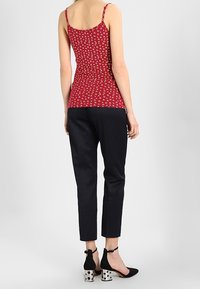 Femme portant un haut sans manches à motifs rouges, un pantalon noir texturé longueur cheville, et des escarpins noirs avec des talons blocs argentés et des brides de cheville.