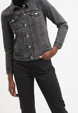 Person trägt verblasste schwarze Jeansjacke mit silbernen Knöpfen und schwarze Jeans, eine Hand in der Tasche, vor weißem Hintergrund.