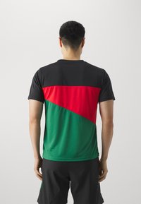 Camisa deportiva para hombre con sección superior negra, parte inferior diagonal en rojo y verde, con cuello redondo y mangas cortas. Textura suave.