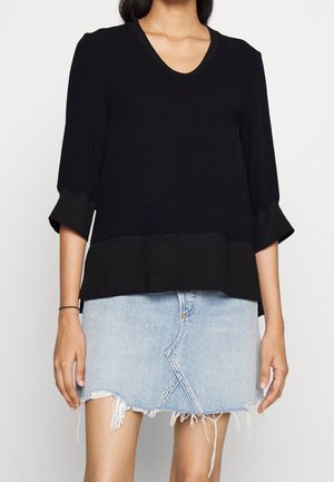 Vrouw draag een zwart top met lange mouwen met omgeslagen mouwen en een lichtblauwe verweerde denim minirok, tegen een effen achtergrond.