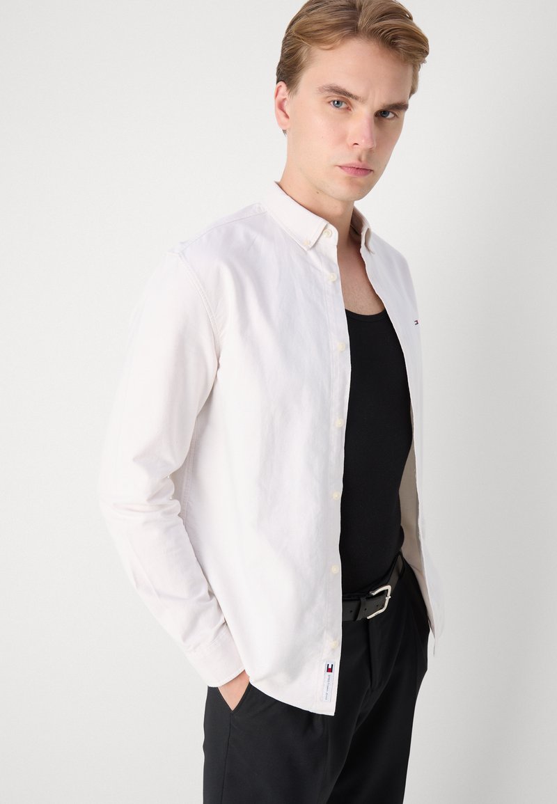 Camisa blanca de manga larga hecha de algodón, con cuello puntiagudo y un pequeño logo en el pecho. Llevada sobre una camiseta sin mangas negra.