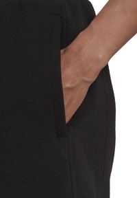 Mano insertada en el bolsillo delantero de pantalones negros, mostrando la textura de la tela y el tono de piel natural.