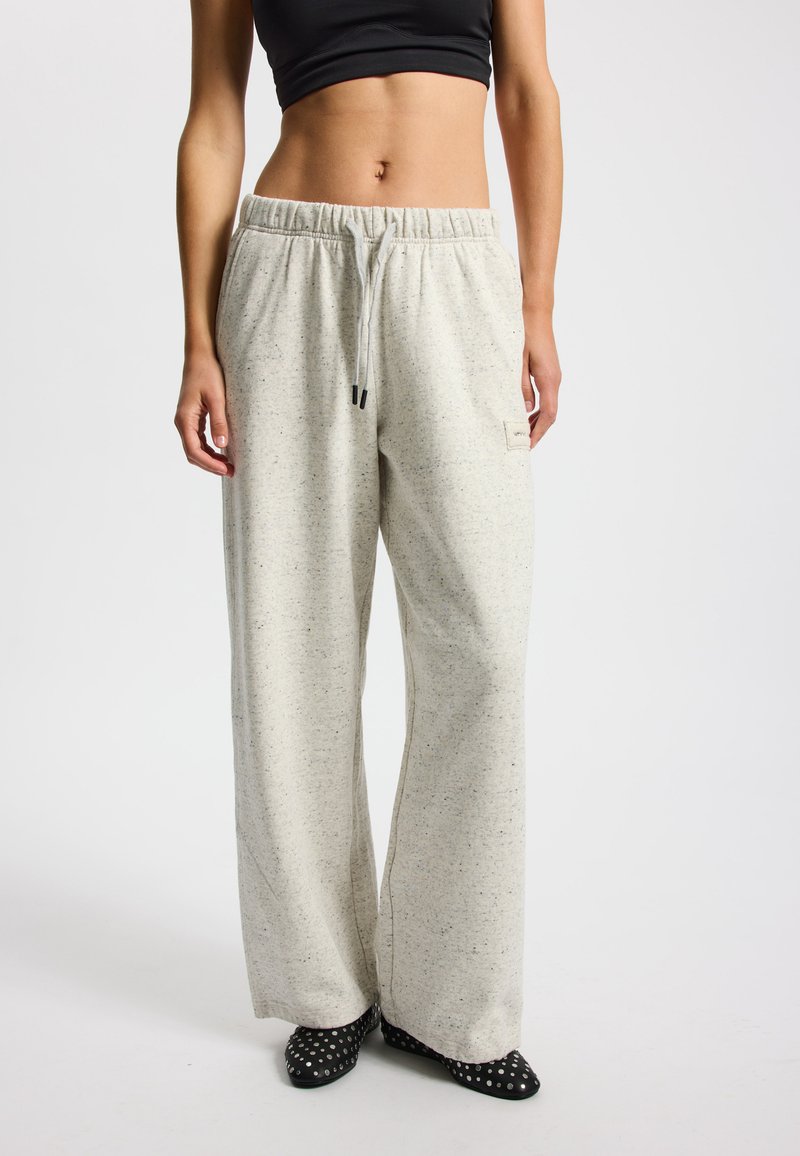 Kobieta ubrana w jasnoszare, szerokie spodnie dresowe z wysokim stanem i sznurkiem, czarny krótki top na ramiączkach oraz czarne, ćwiekowane buty na szpic.