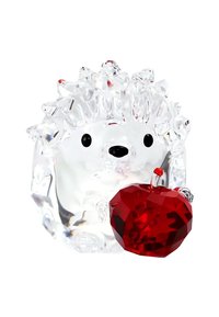 Figura de erizo de cristal transparente sosteniendo una manzana roja facetada contra un fondo blanco.