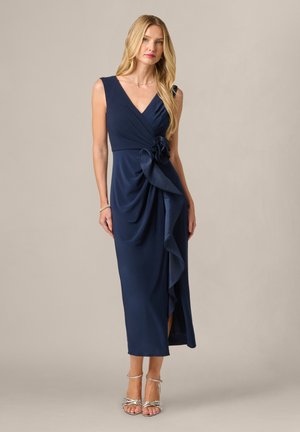 Vrouw met lang blond haar die een mouwloze marineblauwe wikkeljurk met een grote zijstrik en zilveren sandalen met hoge hakken draagt, staand op een beige achtergrond.