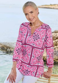 Bunt gemusterte Bluse in Pink und Weiß mit Blumenmotiven; verfügt über einen V-Ausschnitt und Dreiviertelärmel. Getragen mit weißen Hosen.