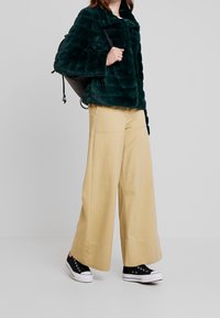 Bunda z umělého kožíšku ve tmavě zelené barvě, s horizontálními pruhy a vysokým límcem, doplněná o světle béžové kalhoty s širokými nohavicemi a černé tenisky.