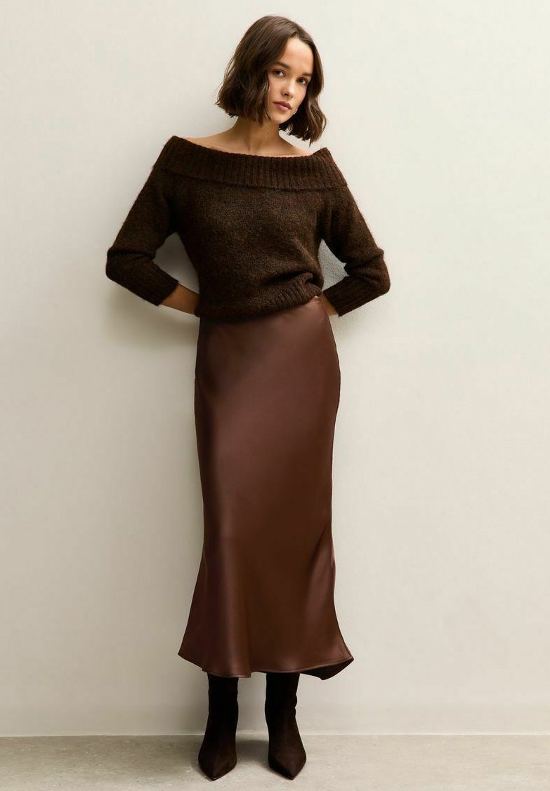 Brown knitted off-shoulder sweater paired with a brown satin maxi skirt and knee-high brown boots, set against a neutral background.
