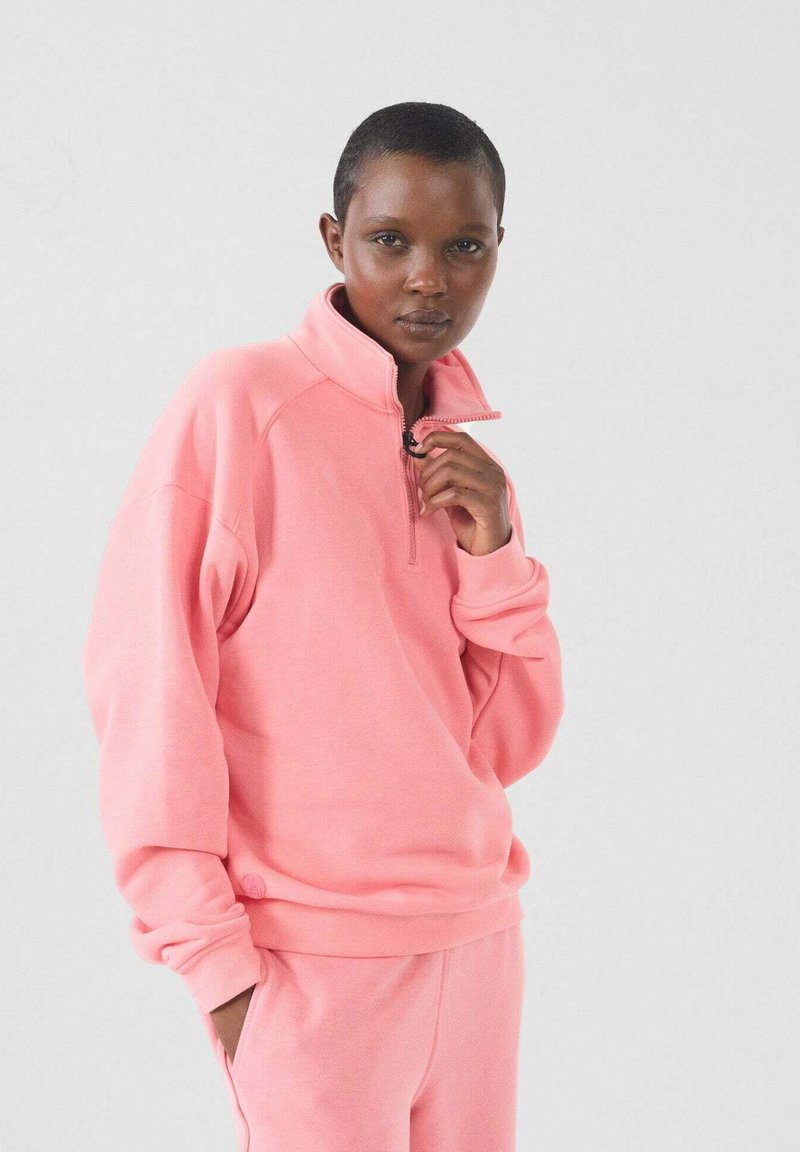 Roze zip-up sweatshirt met een hoge kraag en geribde mouwen. Zachte textuur en relaxte pasvorm, gecombineerd met bijpassende broeken. Eenvoudige ontwerpelementen.