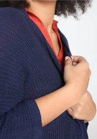 Cárdigan de punto azul marino con superficie texturizada, mangas ajustadas y un frente abierto casual, superpuesto a una camisa de botones roja.