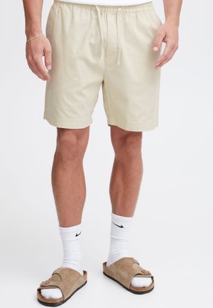 Pantalones cortos beige y ligeros con cintura elástica y cordón. Se llevan con calcetines blancos y sandalias marrones, mostrando un ajuste relajado y un diseño casual.