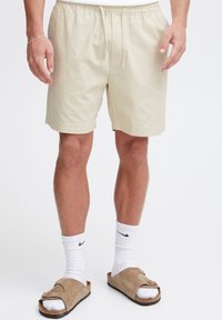 Beige, lightweight shorts with an elastic waistband and drawstring. Worn with white socks and brown sandals, showcasing relaxed fit and casual design.