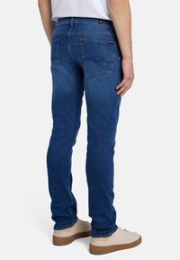 Jeans in denim blu con una vestibilità slim, cinque tasche e cuciture a contrasto. Indossati con scarpe di colore chiaro, mostrando un tessuto strutturato.