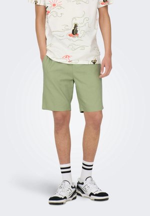 Person iført lysegrønne shorts, hvid T-shirt med farverige tropiske print, hvide sokker med sorte striber og hvide sorte sneakers.