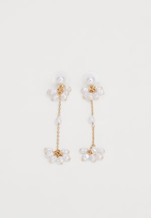 Guldtonede øreringe med kædedetaljer, der har klynger af hvide falske perler. Cirkulær plastikbaggrund for sikker bæring.