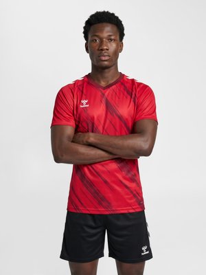 Man stands with arms crossed wearing a red patterned sports shirt and black shorts against a plain white background.